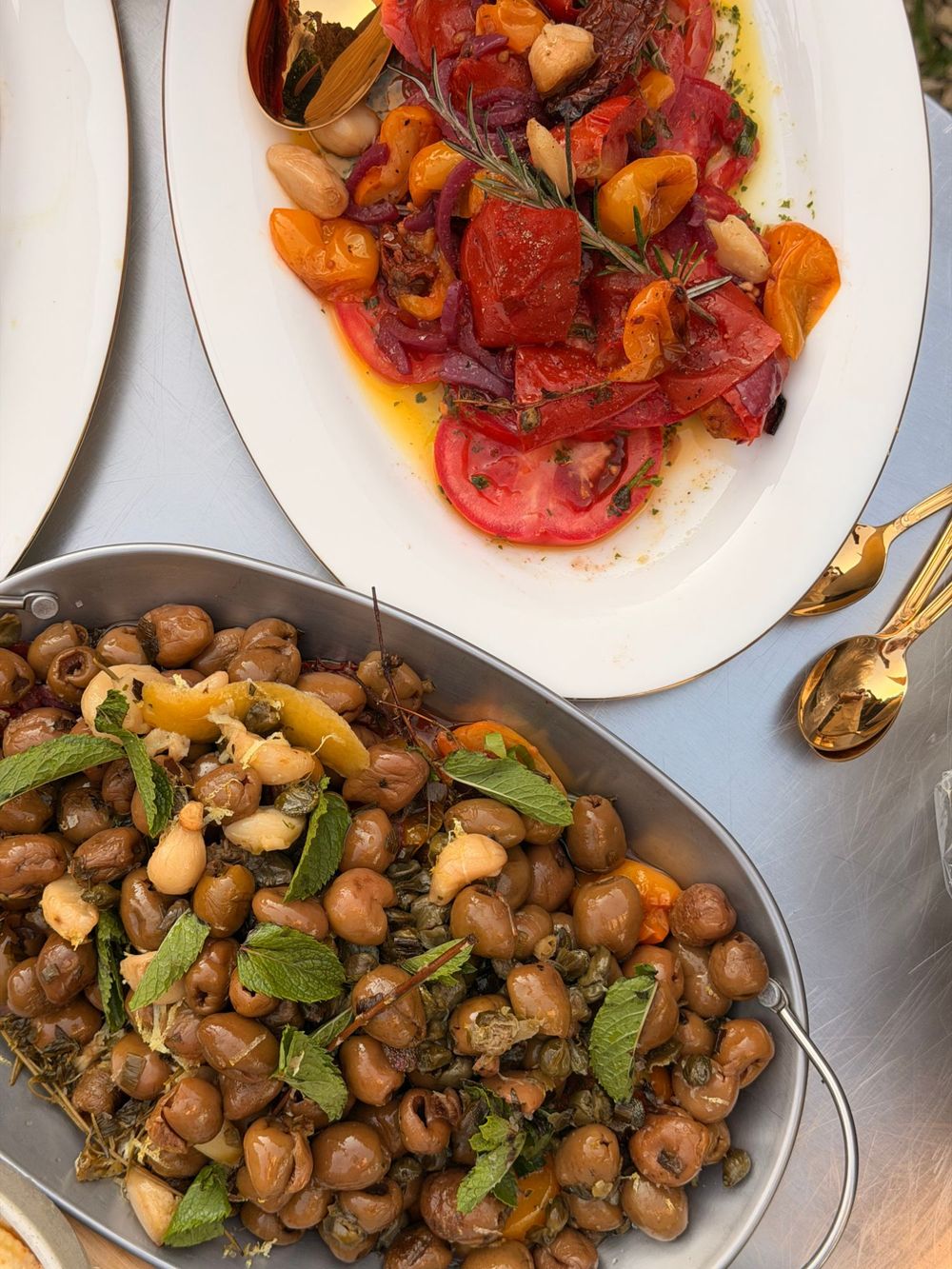 Roasted tomatoes on a white plate and marinated olives in a steel pan, with gold spoons