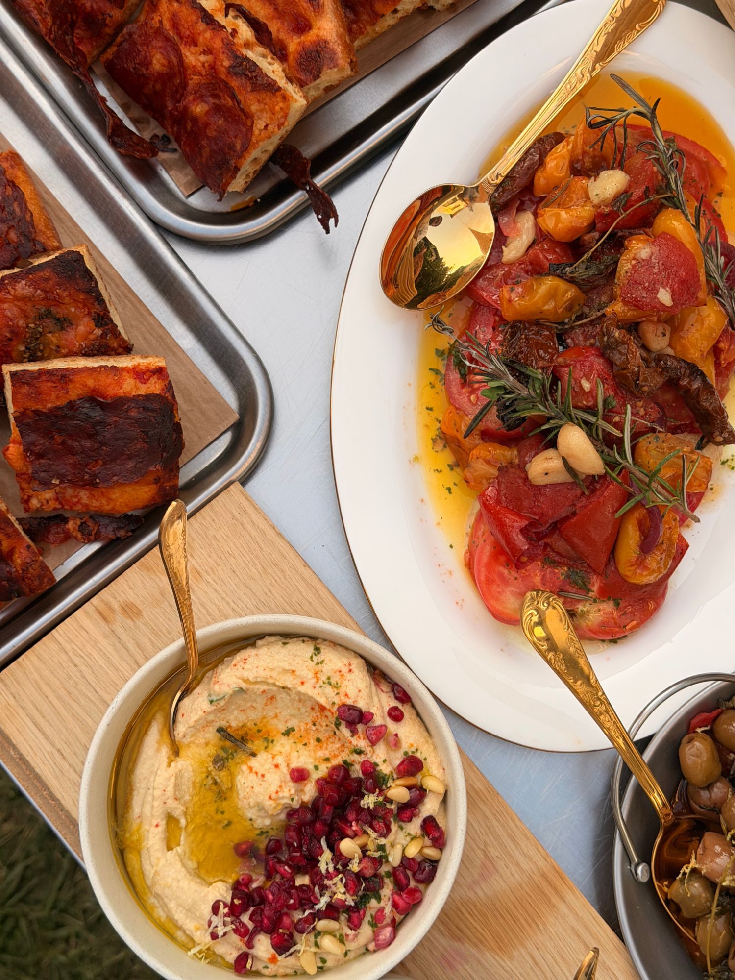 Overhead view of a table: roasted tomatoes, hummus with pomegranate, marinated olives and focaccia, with gold cutlery