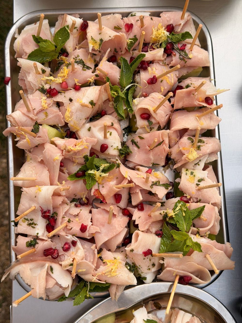 A steel tray of rolled ham skewers with mint, pomegranate seeds and lemon zest